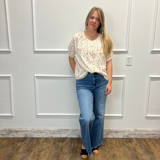 A woman stands in a room with white paneled walls and a wood floor, wearing a cream short-sleeve blouse with a small floral print and blue jeans.