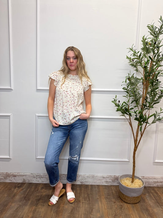 Woman in a floral top and jeans standing next to a plant against a white paneled wall.