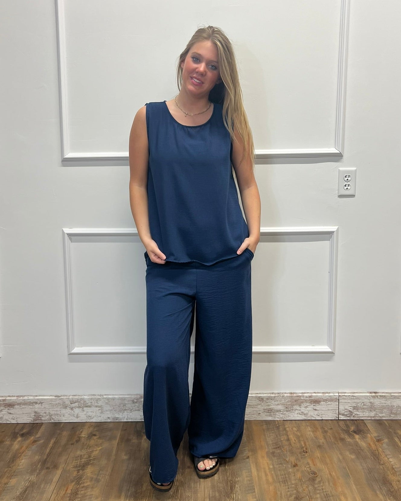 A woman modeling a navy blue sleeveless top and matching wide-leg pants, standing against a white paneled wall with wood flooring.