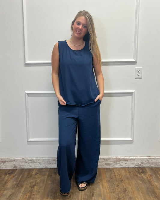 A woman modeling a navy blue sleeveless top and matching wide-leg pants, standing against a white paneled wall with wood flooring.
