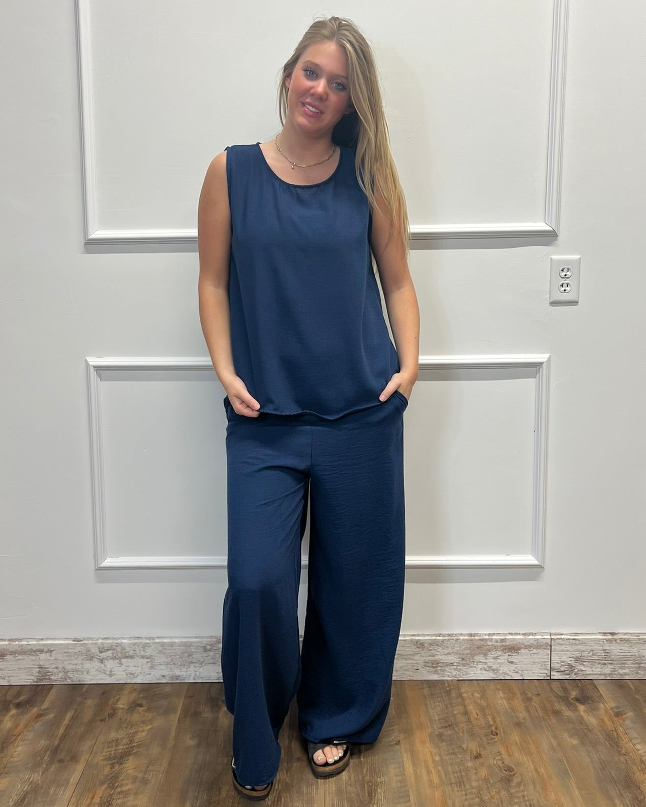 A woman modeling a navy blue sleeveless top and matching wide-leg pants, standing against a white paneled wall with wood flooring.