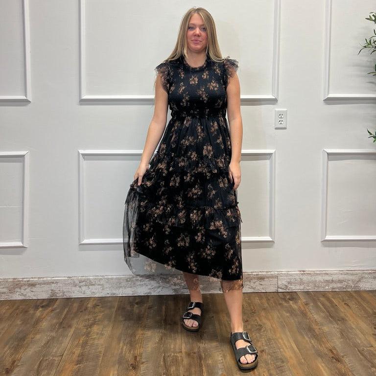 A woman modeling a sleeveless black midi dress with a dark floral pattern and sheer flutter sleeves, standing in front of a white paneled wall on a wood floor.