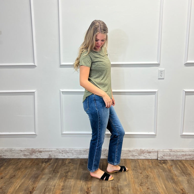 A woman modeling a short-sleeve olive green t-shirt and blue jeans, standing on a wood floor against a white paneled wall.