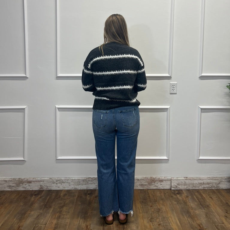 A woman viewed from behind wearing a black knit sweater with white horizontal stripes and blue jeans, standing in front of a white paneled wall on a wood floor.