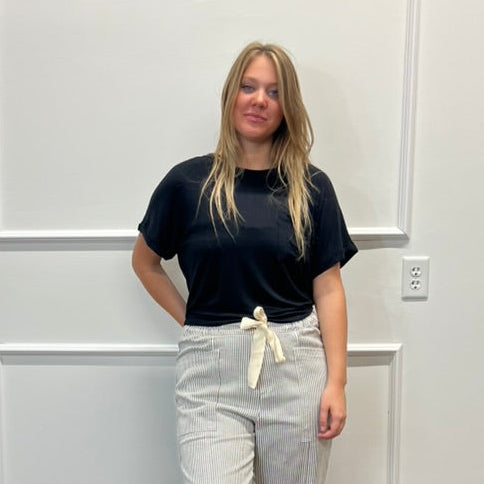 A woman stands in a room with white paneled walls and a wood floor, wearing a black short-sleeve t-shirt, light beige wide-leg pants with a cream tie, and black slide sandals.
