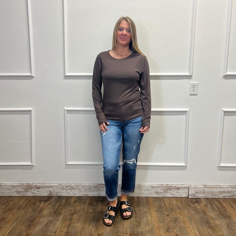A woman stands in a room with white paneled walls and a wood floor, wearing a dark brown long-sleeve crewneck top, blue distressed jeans, and black sandals.