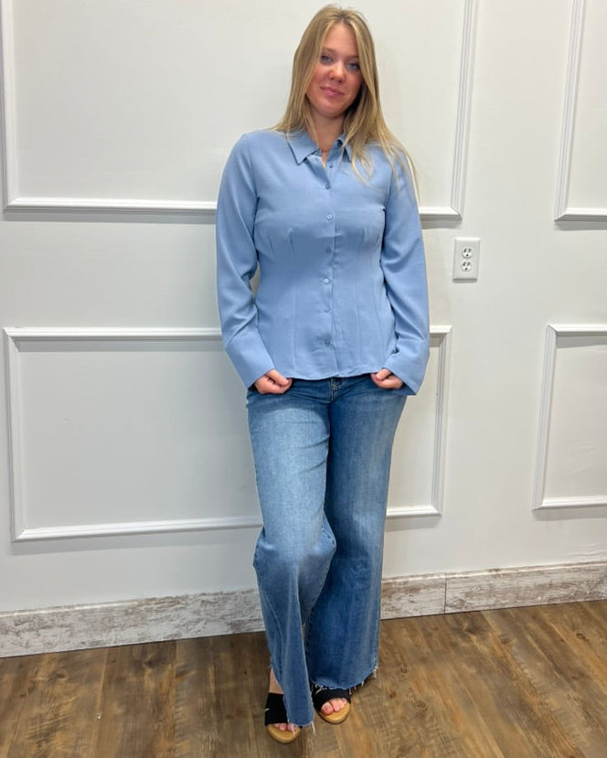 A woman stands against a white paneled wall wearing a light blue long-sleeve button-up shirt and blue jeans, with black sandals on a wood floor.