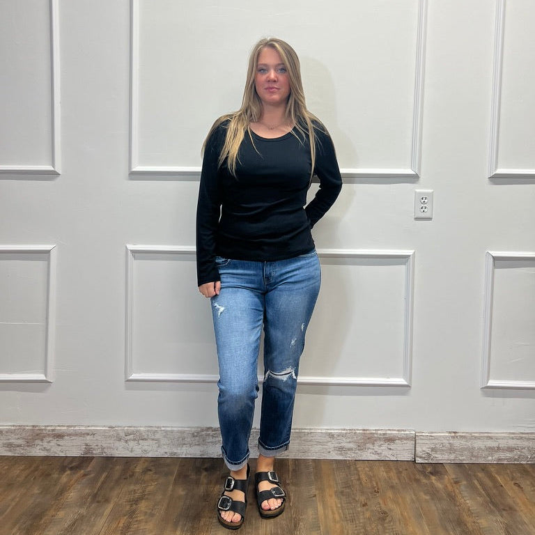 A woman stands in a room with white paneled walls and a wood floor, wearing a black long-sleeve top, blue distressed jeans, and black sandals.
