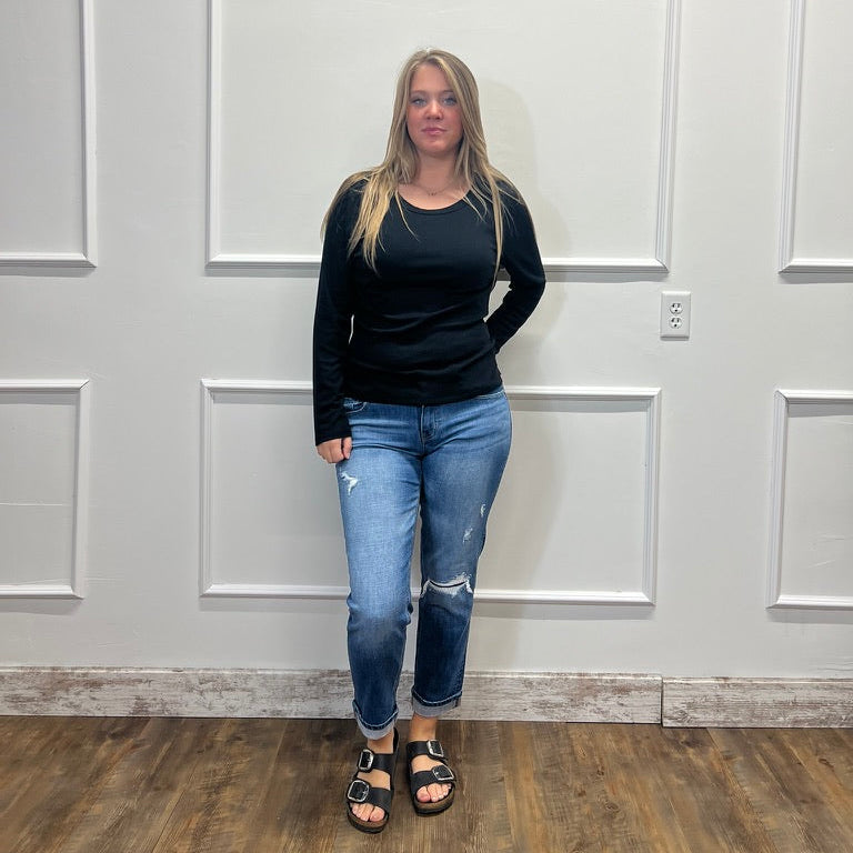 A woman stands in a room with white paneled walls and a wood floor, wearing a black long-sleeve top, blue distressed jeans, and black sandals.