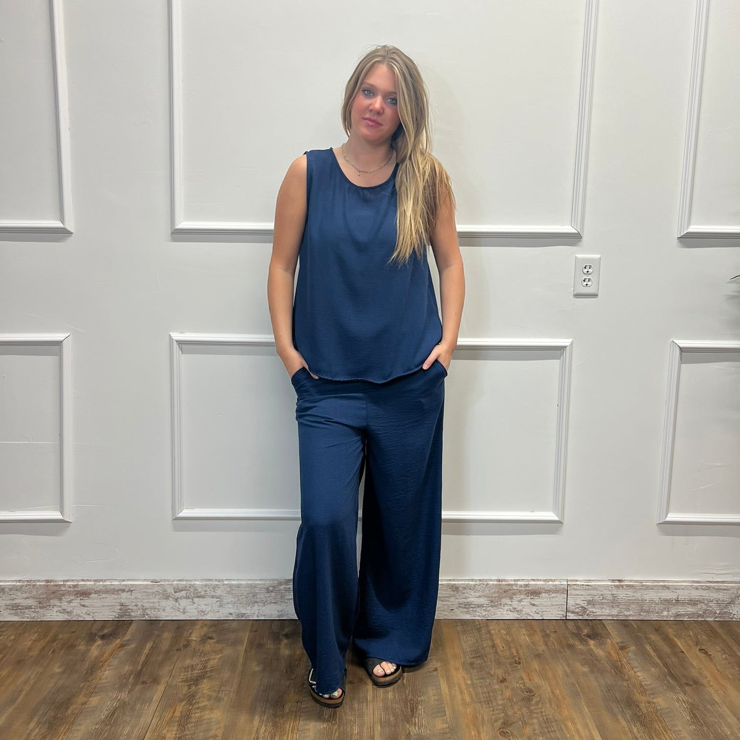 A woman modeling a navy blue sleeveless top and matching wide-leg pants, standing against a white paneled wall on a wood floor.