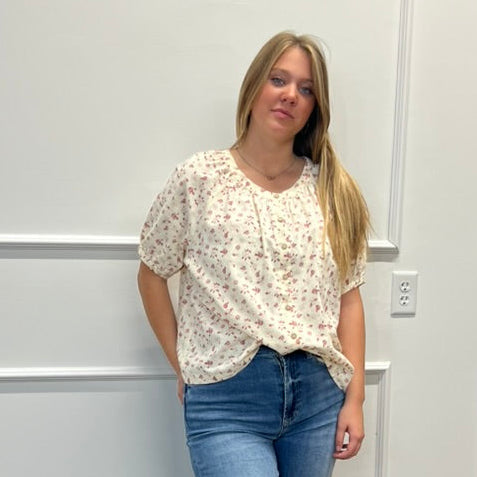 A woman stands indoors against a white paneled wall wearing a cream short-sleeve blouse with a small floral print and blue jeans.
