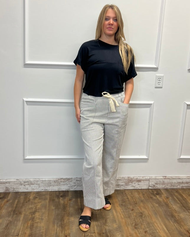 A woman stands indoors against a white paneled wall on a wood floor, wearing a black short-sleeve top, light beige wide-leg drawstring pants, and black slide sandals.