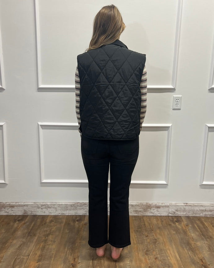 A woman stands with her back to the camera wearing a black diamond-quilted puffer vest over a black and white striped long-sleeve shirt and black pants, standing on a wood floor against a white paneled wall.