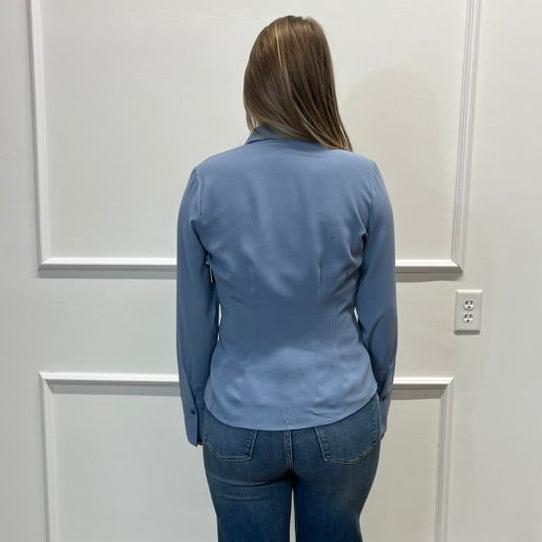 A woman viewed from behind wearing a light blue long-sleeve collared shirt and dark blue wide-leg jeans, standing on a wood floor against a white paneled wall.