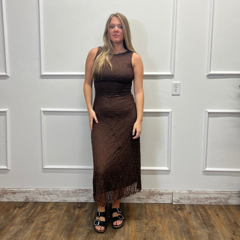 A woman stands in a fitting room wearing a sleeveless, high-neck brown lace dress with a fringe hem and black sandals, set against a white paneled wall and wood floor.