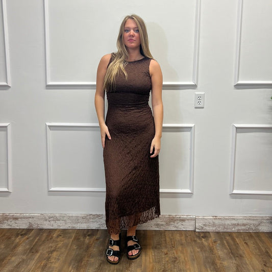 A woman stands in a fitting room wearing a sleeveless, high-neck brown lace dress with a fringe hem and black sandals, set against a white paneled wall and wood floor.