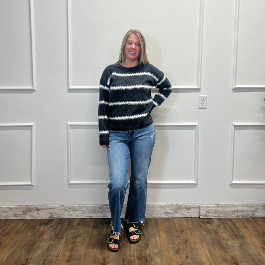 A woman stands in front of a white paneled wall wearing a black and white patterned knit sweater, blue jeans, and black sandals.
