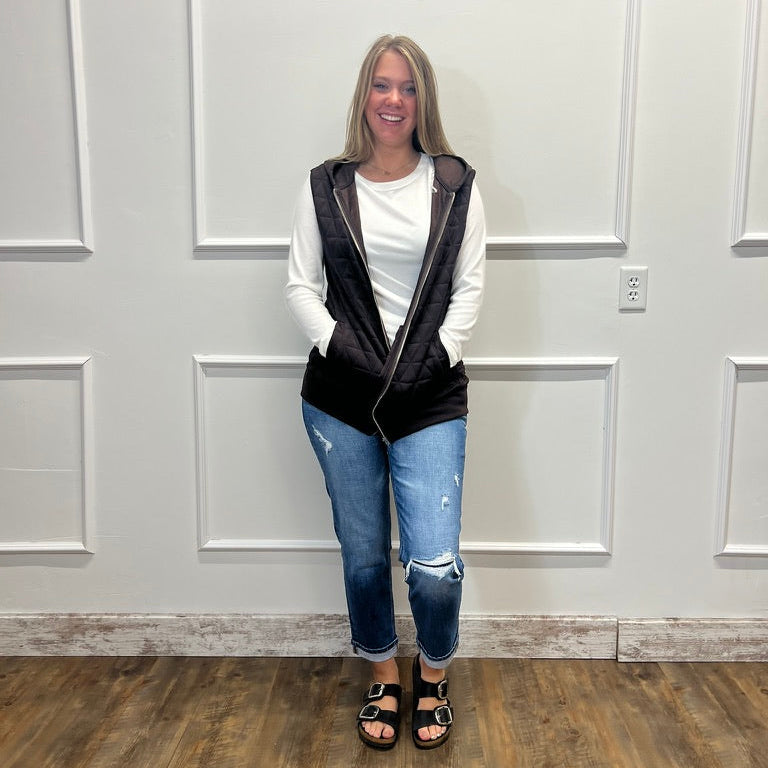 A woman stands indoors against a white paneled wall wearing a black zip-front vest over a white long-sleeve top, blue ripped jeans, and black sandals.