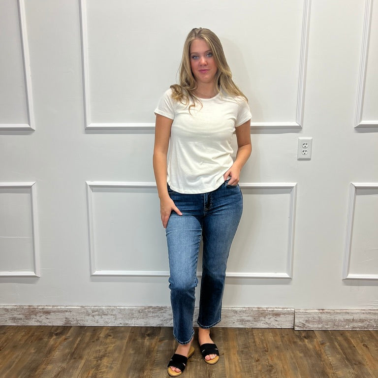 A woman modeling a white short-sleeve t-shirt and blue jeans, standing against a white paneled wall on a wood-look floor.