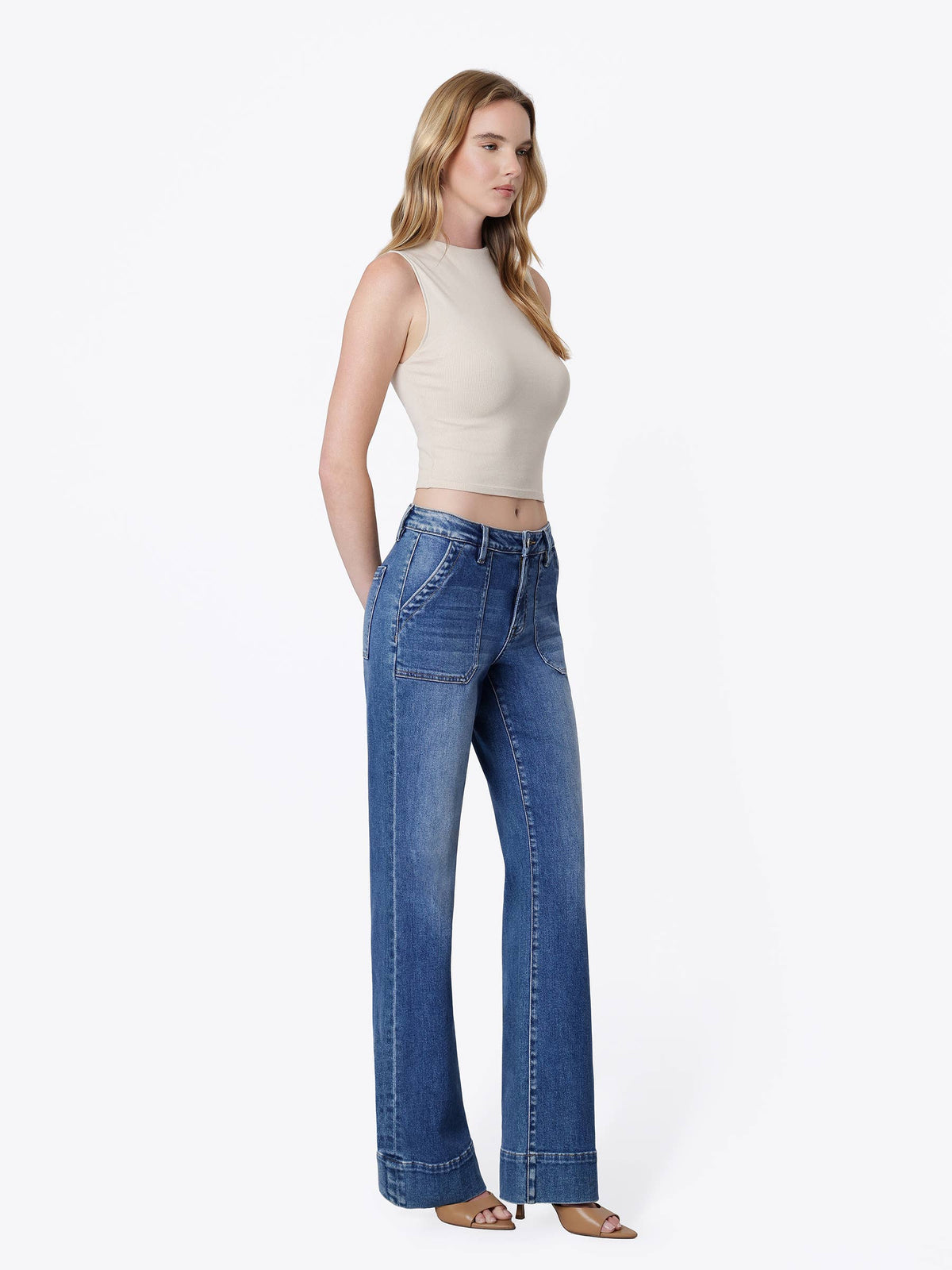 A woman modeling a pair of high-waisted, wide-leg blue jeans with a beige sleeveless crop top, standing against a plain white studio background.