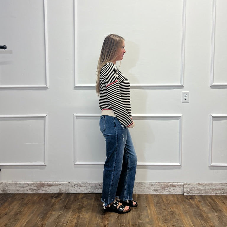 A woman stands in profile wearing a black and white striped long-sleeve crop top, blue jeans, and black sandals, standing against a white paneled wall with wood flooring.