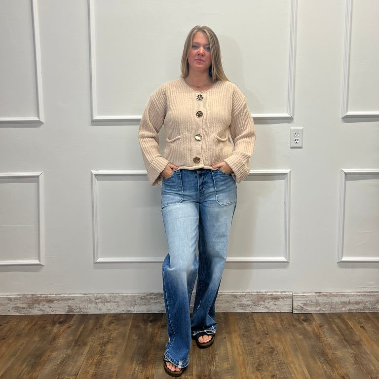 A woman stands in a room with white paneled walls and a wood floor, wearing a beige ribbed cardigan with large buttons and blue jeans.