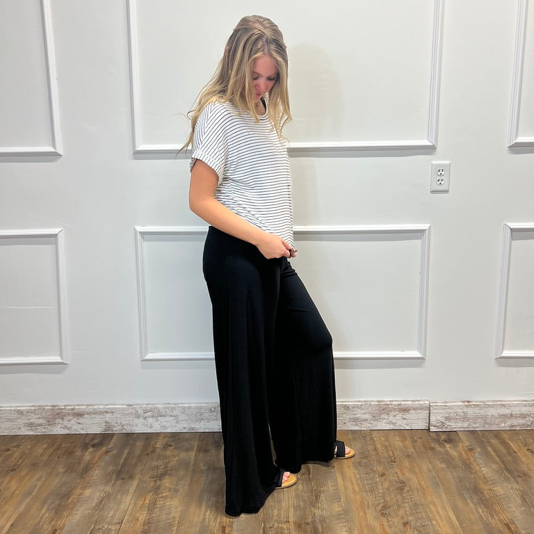 A woman stands in a room with white paneled walls and a wood floor, wearing a white short-sleeve t-shirt with thin black horizontal stripes and black wide-leg pants.