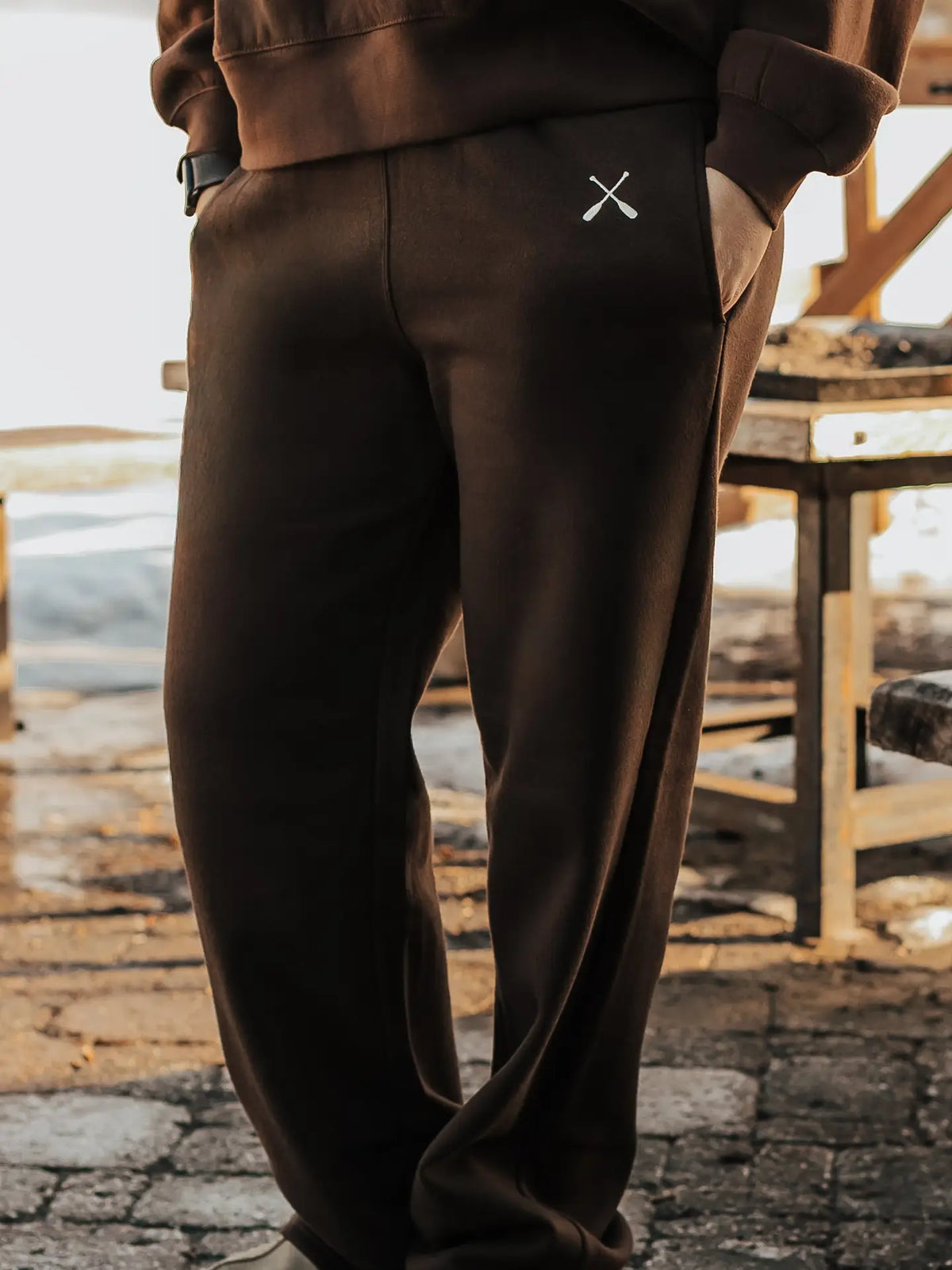 A person wearing dark brown sweatpants with a small white crossed-objects logo on the left hip, paired with a matching brown top, standing outdoors on a stone surface.