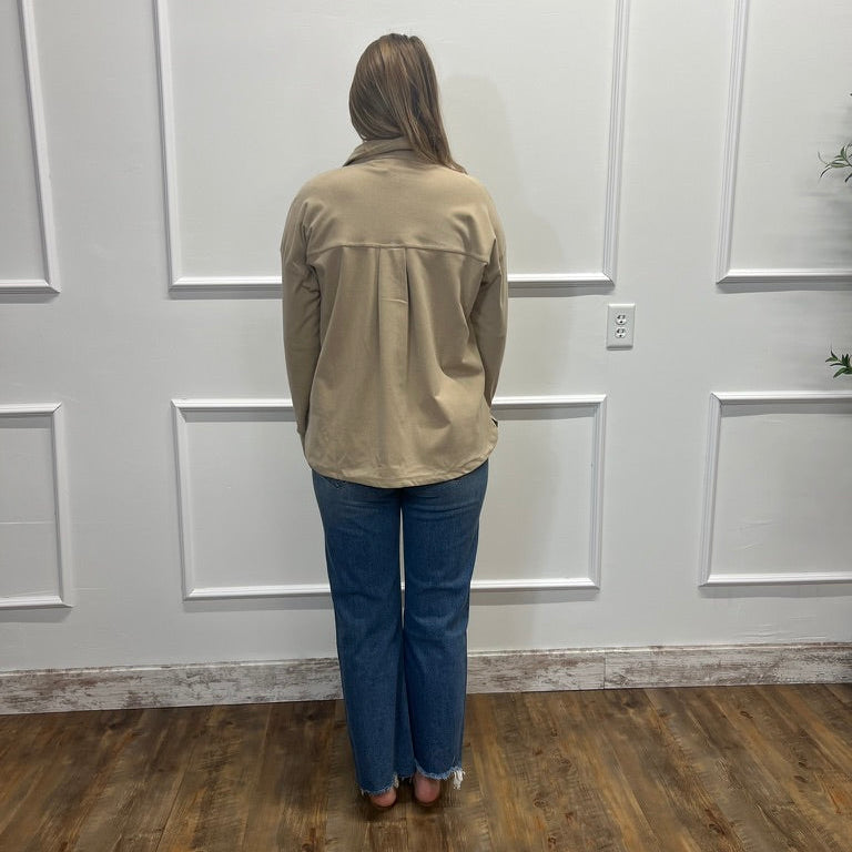 A woman stands with her back to the camera wearing a beige utility-style jacket and blue jeans in a room with white paneled walls and a wood floor.
