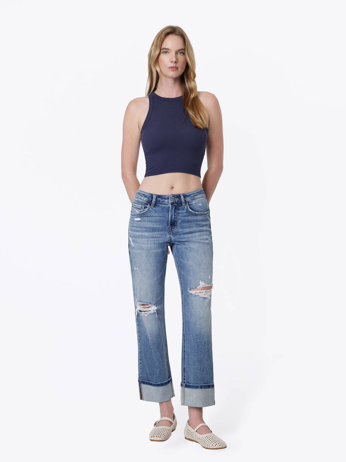 A woman modeling a navy blue halter crop top and light-wash blue distressed jeans with rolled cuffs, paired with beige flat shoes, against a plain white studio background.