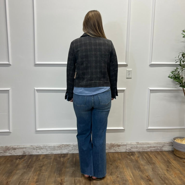 A woman stands with her back to the camera wearing a dark gray and black plaid flannel shirt jacket, a light blue t-shirt, and blue flare jeans, standing in front of a white paneled wall on a wood floor.
