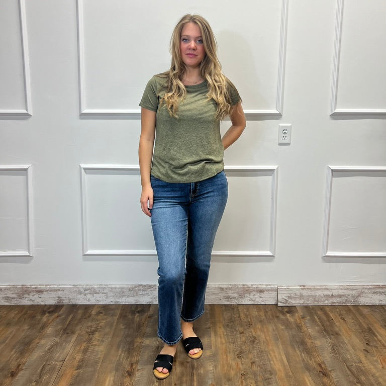 A woman modeling a short-sleeve olive green t-shirt and blue jeans, standing in front of a white paneled wall on a wood floor.
