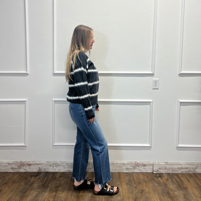 A woman stands in profile wearing a black and white horizontally striped knit sweater, blue jeans, and black slide sandals, standing against a white paneled wall on a wood floor.