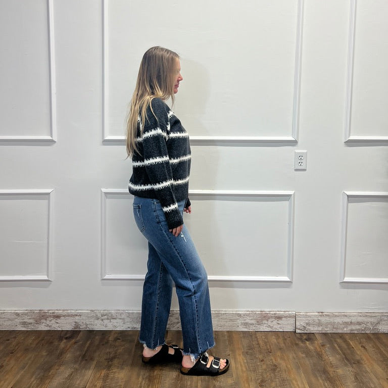 A woman stands in profile wearing a black and white horizontally striped knit sweater, blue jeans, and black slide sandals, standing against a white paneled wall on a wood floor.
