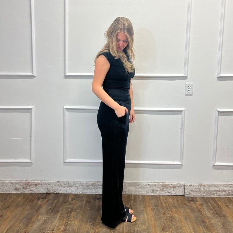 A woman with blonde hair stands in profile wearing a sleeveless black maxi dress with side pockets and black heeled sandals, posing in front of a white paneled wall on a wood floor.