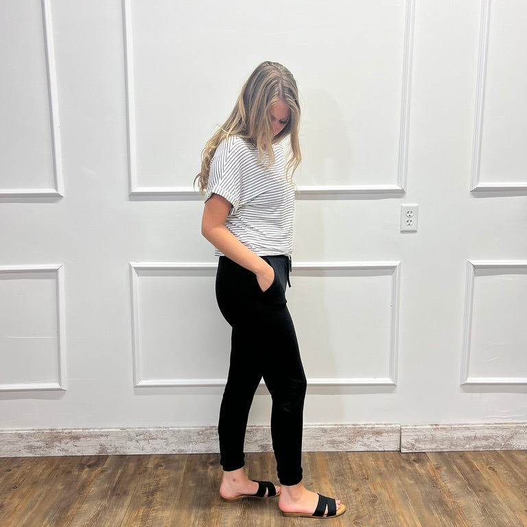 A woman stands in profile wearing a white short-sleeve t-shirt with thin black horizontal stripes, black high-waisted leggings, and black slide sandals, set against a white paneled wall and a wood-look floor.
