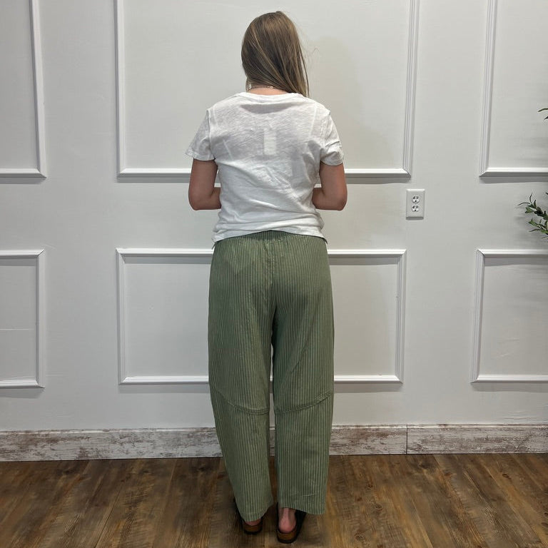A woman viewed from behind wearing a white t-shirt and olive green wide-leg pleated pants, standing in front of a white paneled wall on a wood floor.