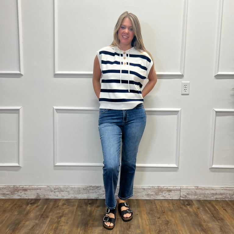 A woman stands against a white paneled wall wearing a sleeveless navy and white striped hoodie with blue jeans and black sandals.
