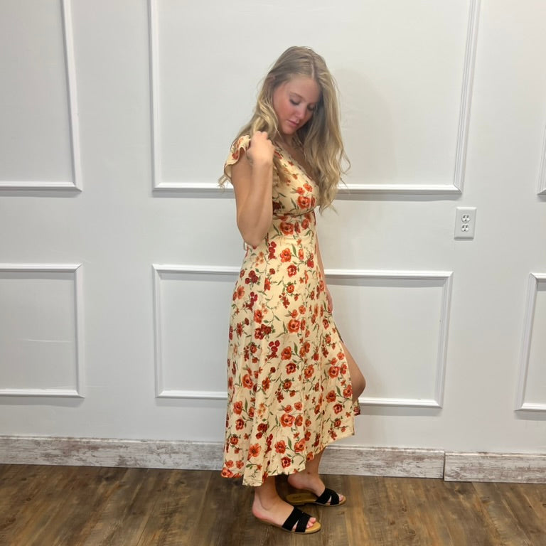 A woman modeling a sleeveless, knee-length cream dress with a red and orange floral pattern, standing on a wood floor against a white paneled wall.