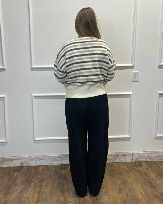 A woman stands with her back to the camera wearing a cream and gray striped long-sleeve knit top and black wide-leg pants, standing on a wood floor against a white paneled wall.