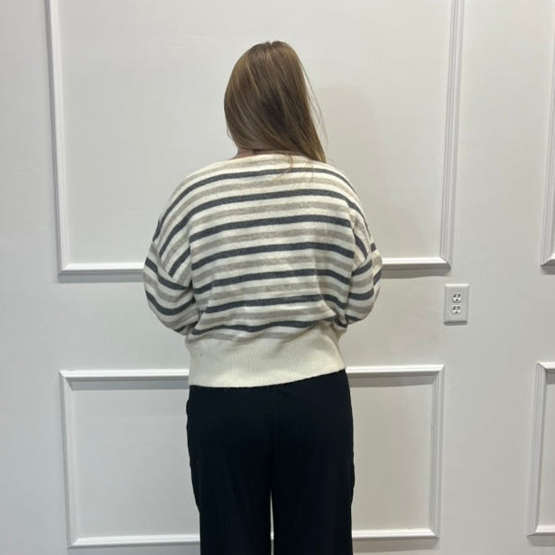 A woman stands with her back to the camera wearing a cream and gray striped long-sleeve knit top and black wide-leg pants, standing on a wood floor against a white paneled wall.