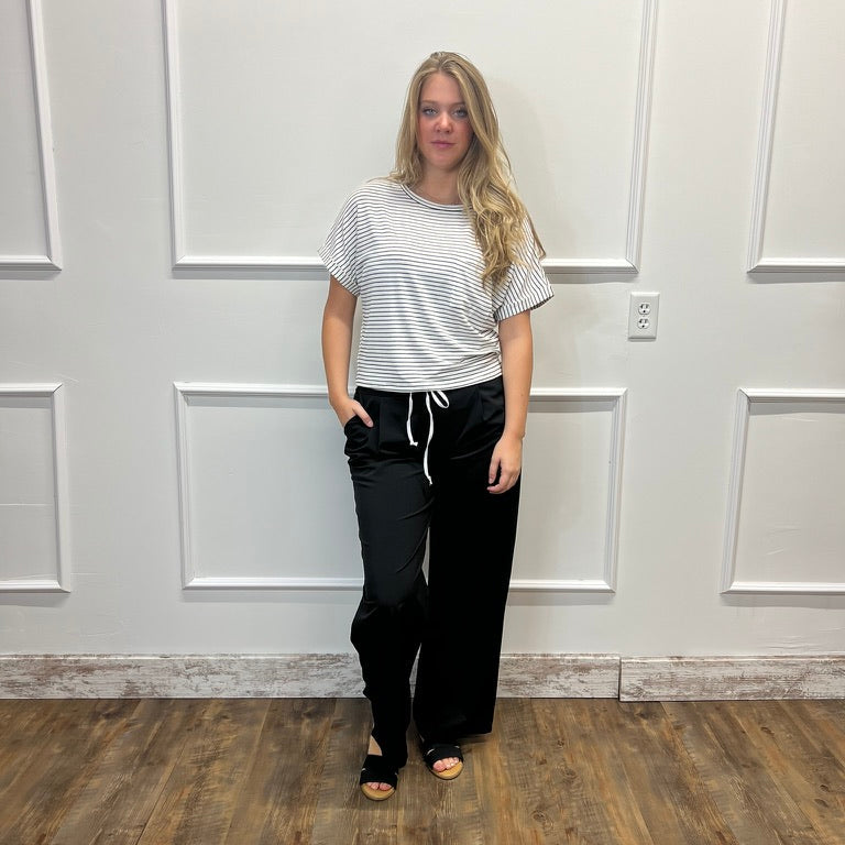 A woman stands in a room with white paneled walls and a wood floor, wearing a white short-sleeve t-shirt with thin black horizontal stripes and black wide-leg drawstring pants.
