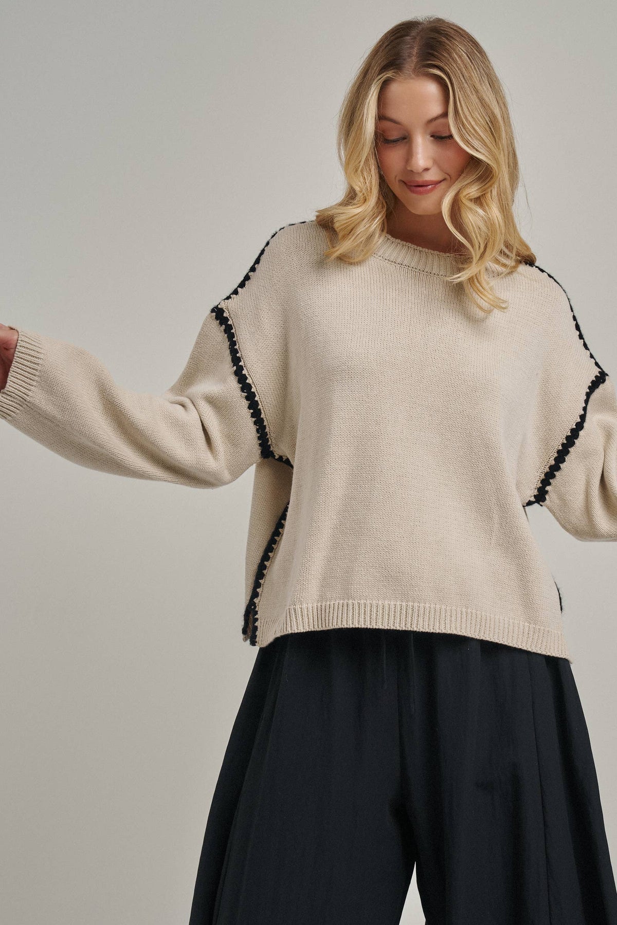 A woman wearing a cream-colored knit sweater with black pom-pom trim along the shoulders and sides, paired with dark wide-leg pants, against a plain light gray studio background.