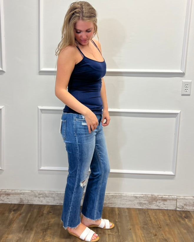 A woman modeling a dark navy blue tank top and blue jeans with white slide sandals, standing against a white paneled wall on a wood floor.