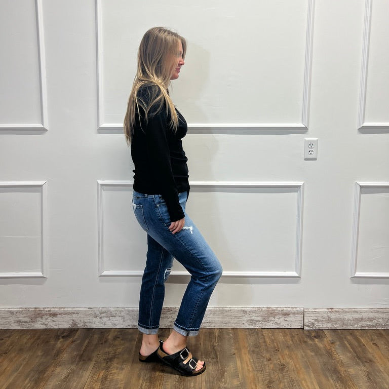 A woman stands in profile wearing a black long-sleeve top, blue cuffed jeans, and black slide sandals with gold hardware, set against a white paneled wall and wood floor.