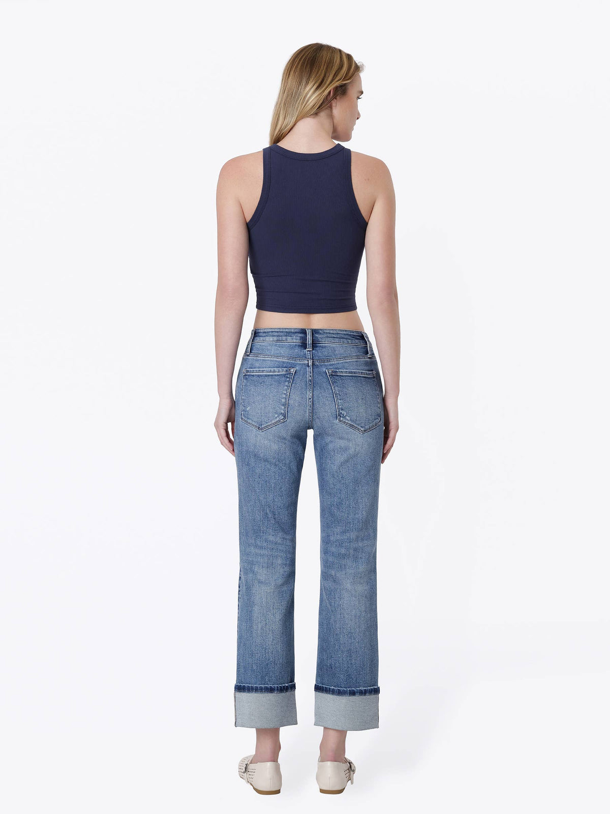 A woman shown from the back wearing a navy blue ribbed crop top and light blue high-waisted straight-leg jeans with rolled cuffs, paired with beige flat shoes, against a plain white studio background.