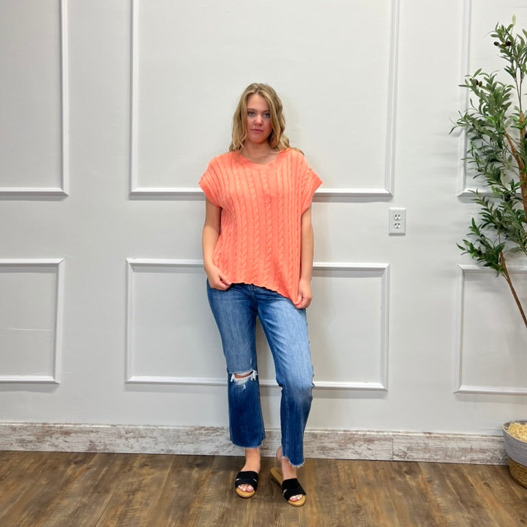 Woman wearing a coral top and blue jeans standing in a room with white paneled walls and a plant.