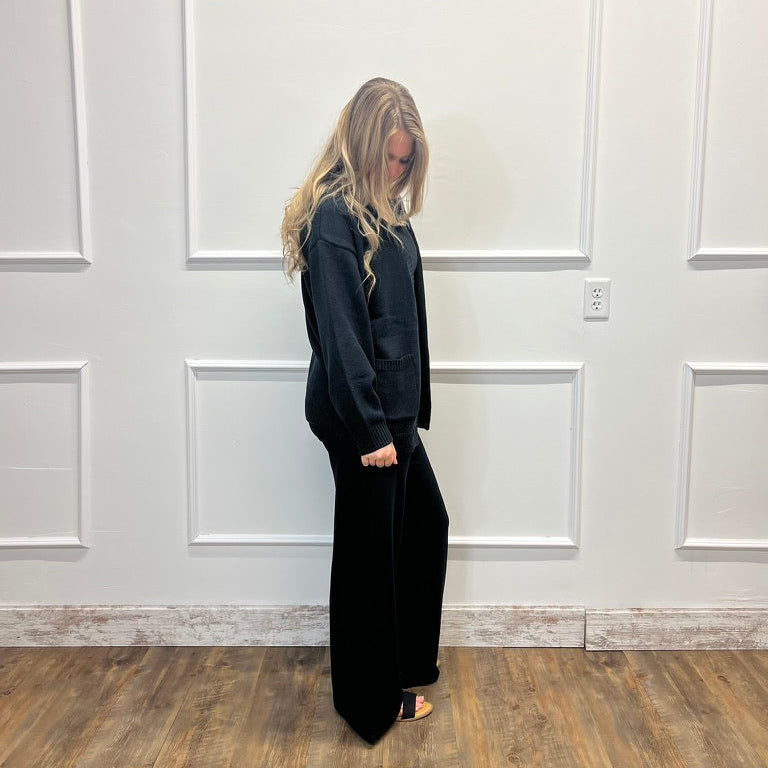 A woman with blonde hair stands in profile wearing a black long-sleeve top and black wide-leg pants against a white paneled wall with a wood floor.