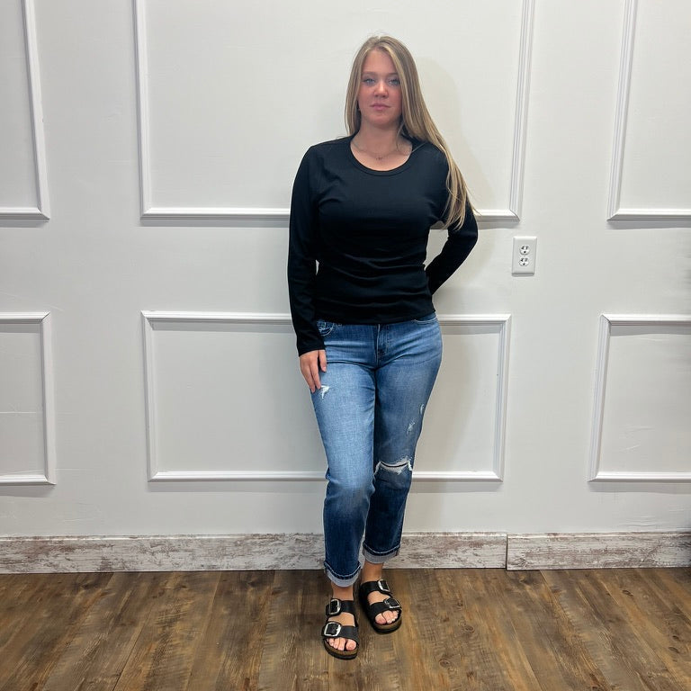 A woman stands in a room with white paneled walls and a wood floor, wearing a black long-sleeve crewneck top, blue ripped jeans, and black slide sandals.