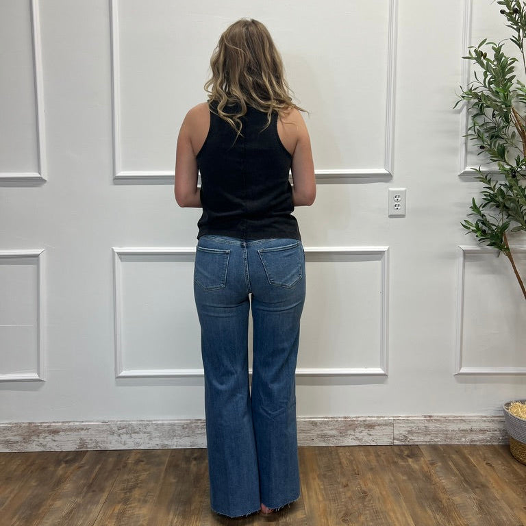 Person wearing blue jeans and a black top standing in a room with white paneled walls and wooden flooring.